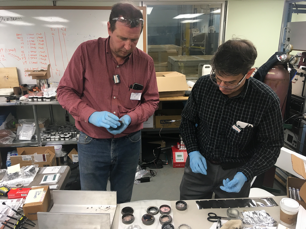 Two men standing in a cluttered room and wearing blue latex gloves examine samples in small disk-shaped containers.