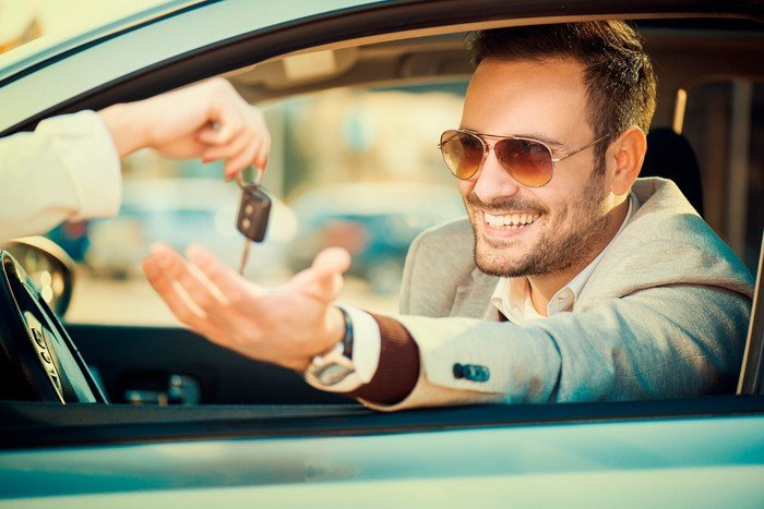 Une personne souriante assise dans sa voiture, tenant sa main par la fenêtre pour retirer les clés d'une autre personne.
