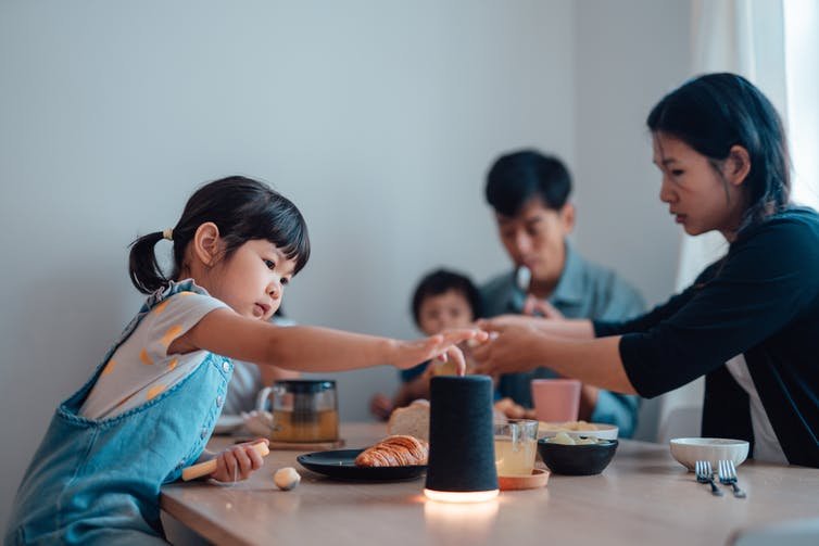 un tout-petit touche le haut d'un cylindre noir sur une table à manger pendant qu'une famille mange en arrière-plan