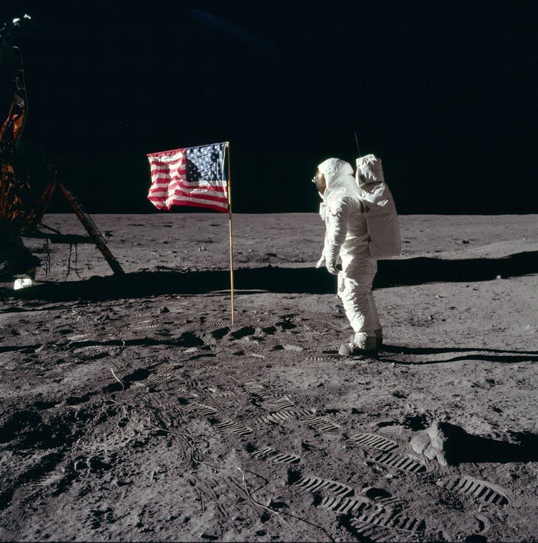Buzz Aldrin en combinaison spatiale à la surface de la Lune à côté du drapeau américain.