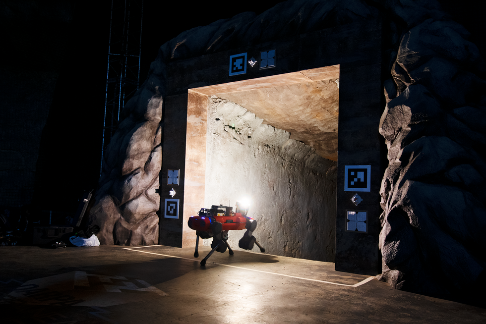 A photo of a dark area with a quad legged robot lighting up a shaft of stone.