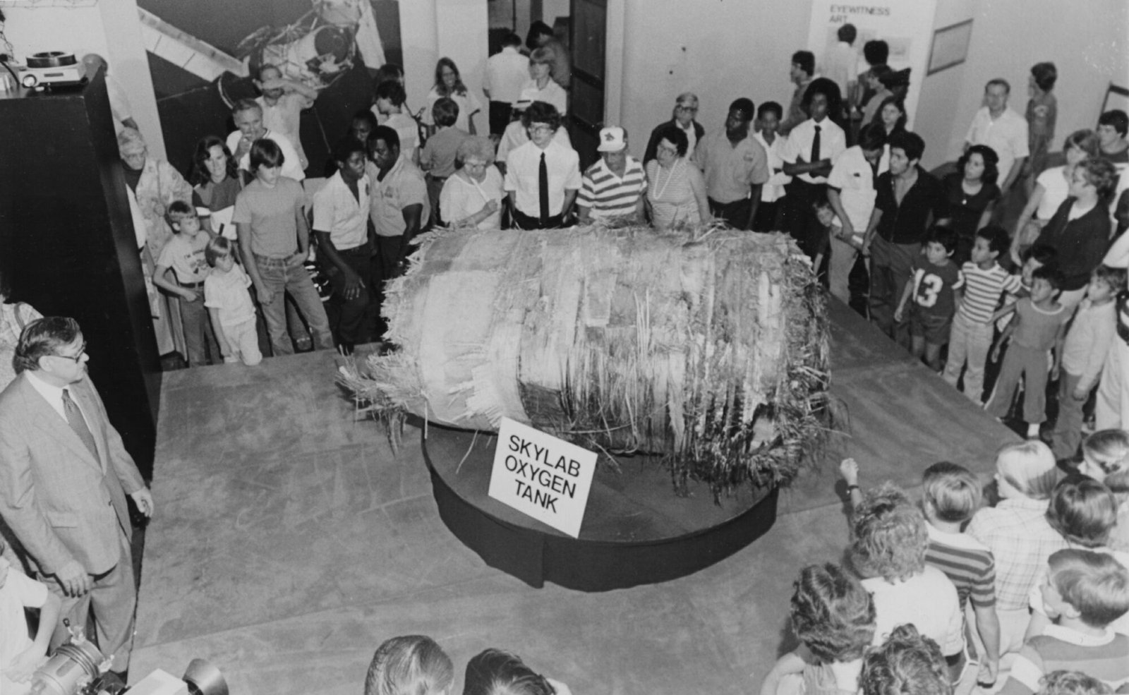 Une photo en noir et blanc de 1979. Un groupe de visiteurs du musée se rassemble en cercle autour d'un gros morceau de métal déchiqueté en forme de cylindre. Il repose sur un piédestal rond avec un panneau indiquant