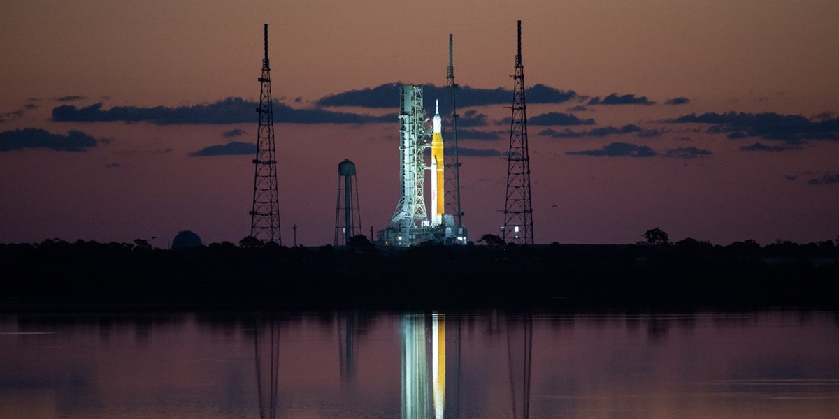 La scène de lancement Artemis I est illuminée à l'aube au Kennedy Space Center en Floride.  Le vaisseau spatial Orion blanc de la NASA est fixé au sommet de la grande fusée cylindrique orange et blanche du système de lancement spatial.