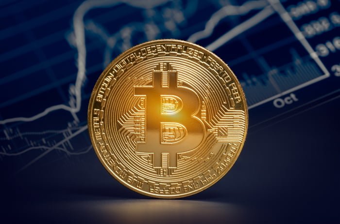 Un Bitcoin en or physique se tenait sur le côté devant un tableau des prix des crypto-monnaies avec volume.