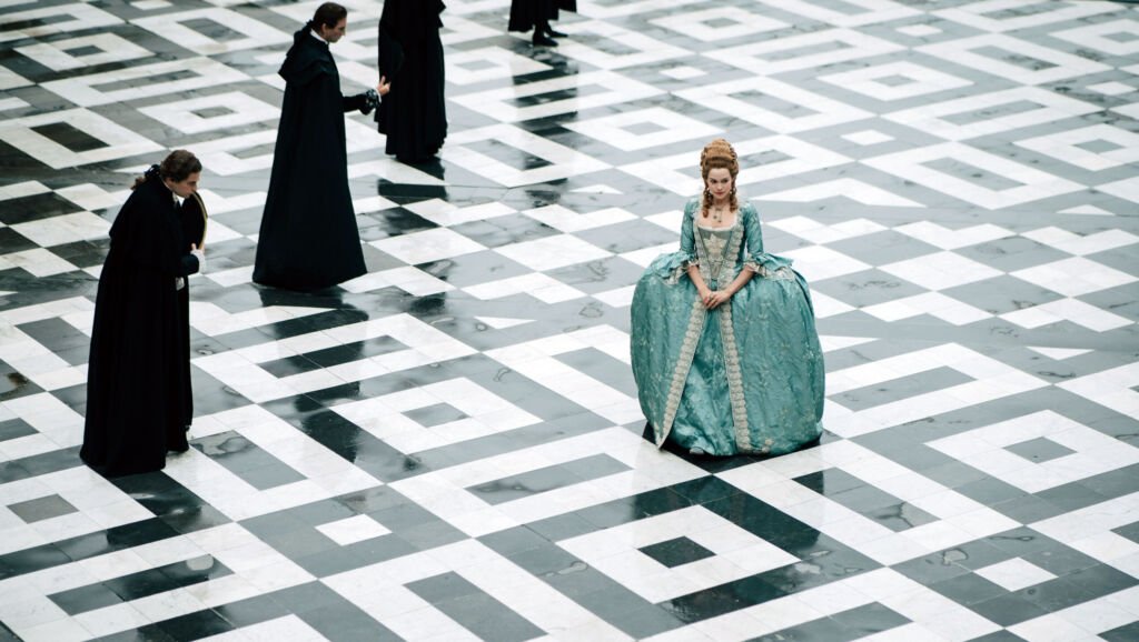 Une vue aérienne d'un sol de palais brillant avec des motifs géométriques en noir et blanc, où une femme se tient debout, les mains jointes et reposant sur sa robe à jupe large. Des silhouettes en robes noires se tiennent en ligne à sa gauche et s'inclinent légèrement.
