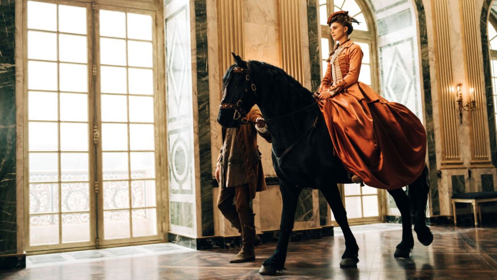 Une femme vêtue d'une longue robe corail brillante est assise en selle sur un cheval qui est conduit dans un couloir du palais avec de hautes fenêtres cintrées.