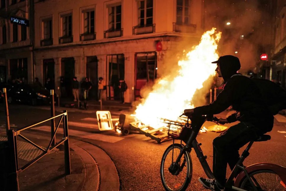 TOPSHOT - Un résident local passe à vélo devant un incendie lors d'une manifestation après que le gouvernement français a fait adopter une réforme des retraites par le parlement sans vote, en utilisant l'article 49,3 de la constitution, à Lyon le 16 mars 2023. - Le président français le 16 mars, une réforme controversée des retraites a été adoptée sans vote par le parlement, déployant un pouvoir constitutionnel rarement utilisé qui risque d'enflammer les protestations. Cette décision était un aveu que son gouvernement n'avait pas la majorité à l'Assemblée nationale pour adopter la législation visant à relever l'âge de la retraite de 62 à 64 ans. (Photo par OLIVIER CHASSIGNOLE / AFP) (Photo par OLIVIER CHASSIGNOLE/AFP via Getty Images)