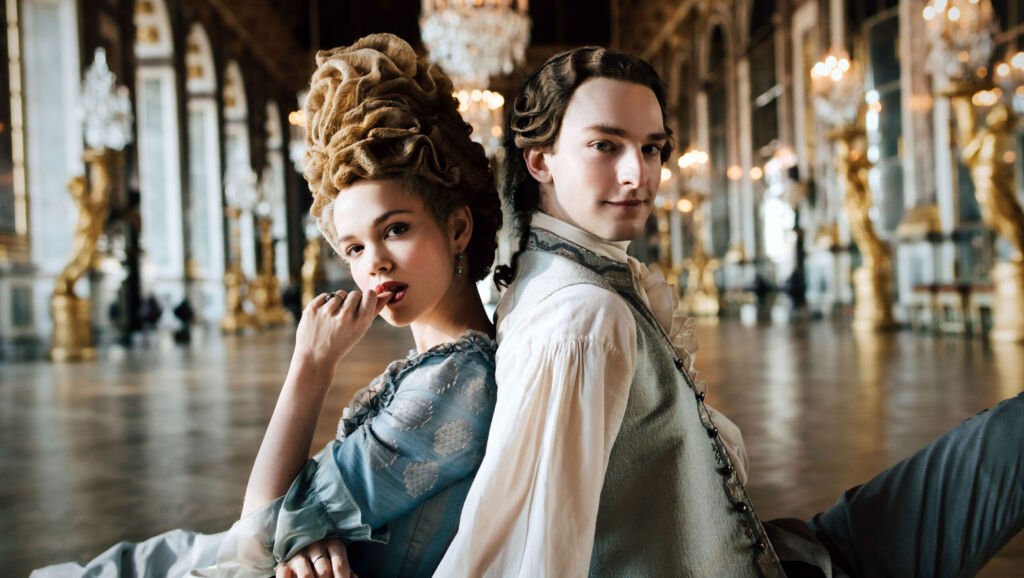 Un jeune homme et une femme s'assoient dans des vêtements formels sur le sol dos à dos et regardent la caméra. Un lustre et de hautes fenêtres sont visibles en arrière-plan.