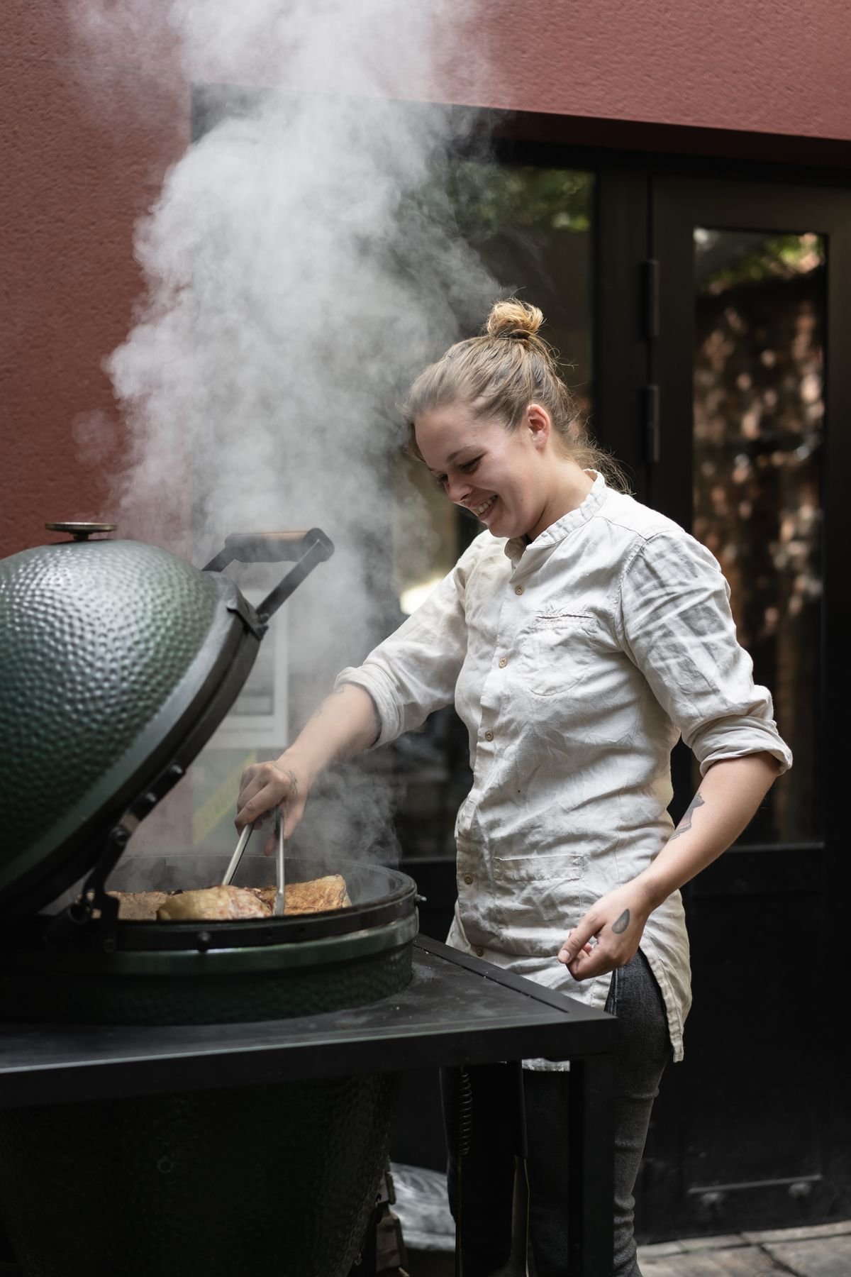 A chef cooks on an outdoor grill, which releases smoke. 