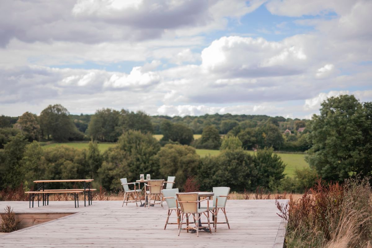 Patio tables on a long wooden deck set in front of rolling hills and trees on a cloudy day. 