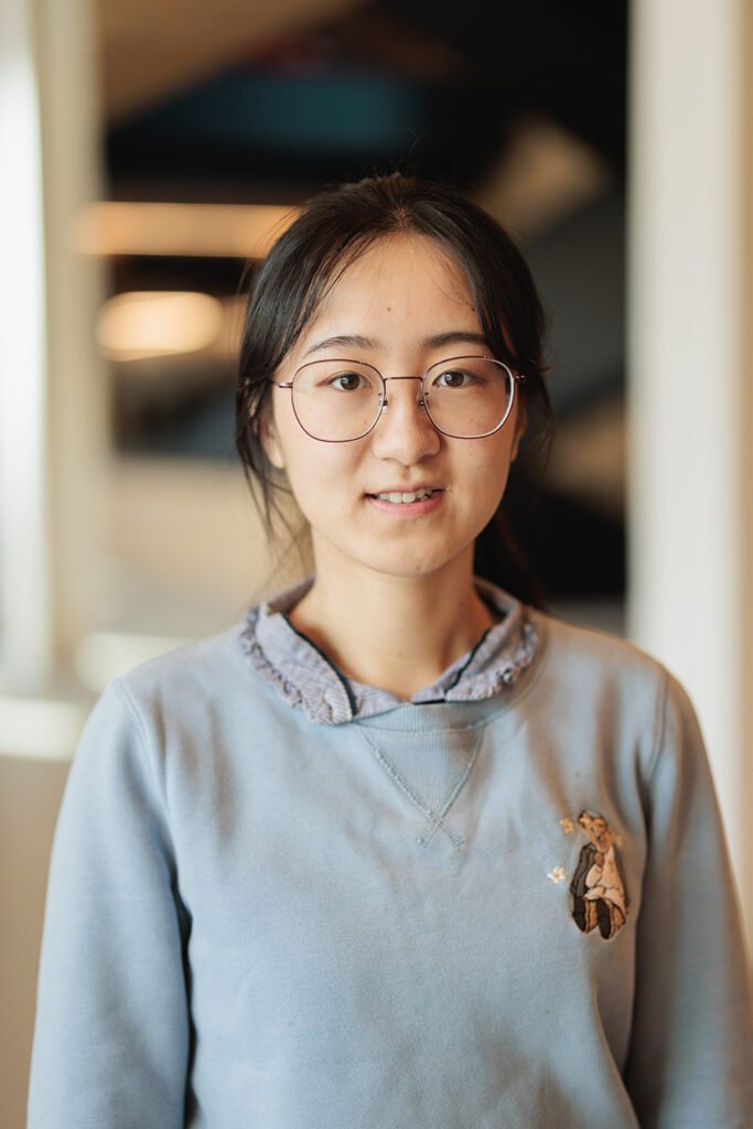 Photo : Yile Wang, une jeune femme asiatique portant des lunettes filaires rondes et un sweat-shirt bleu-gris clair, sourit et pose pour la caméra.
