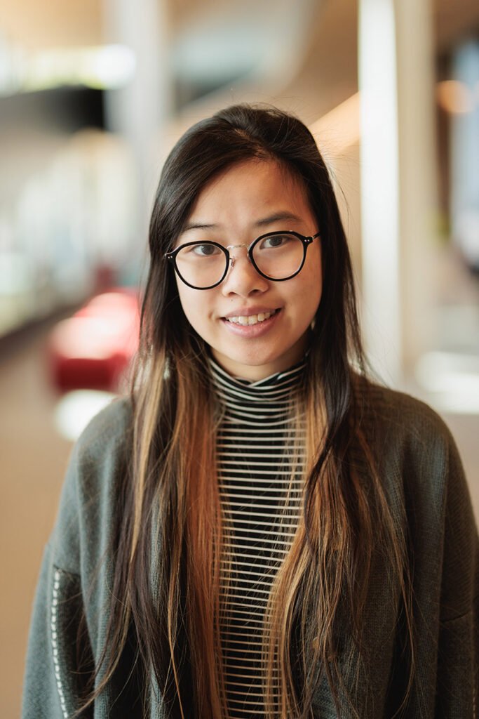 Photo : Samantha Chen, une jeune femme asiatique aux longs cheveux brun foncé avec des reflets brun clair et portant des lunettes noires, un col roulé à rayures noires et blanches et un cardigan gris foncé, sourit et pose pour la caméra.