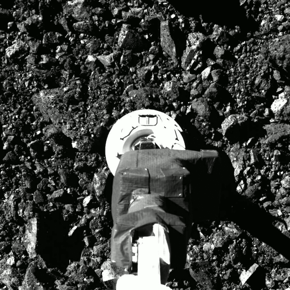 Cette image animée, en noir et blanc, montre une surface déchiquetée recouverte de rochers. Un bras métallique, éclairé par la lumière, avec un dispositif métallique rond en bas, tape brièvement la surface et recule rapidement.
