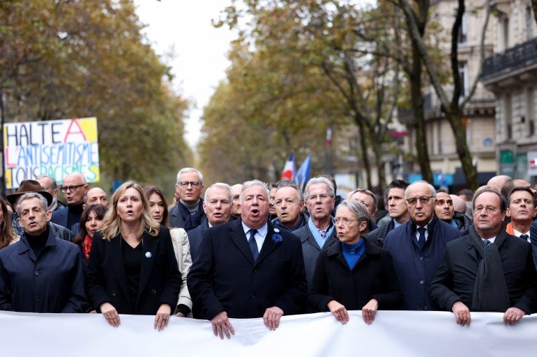 De hauts responsables politiques français, dont la présidente de l'Assemblée nationale Yael Braun-Pivet, la Première ministre Elisabeth Borne, le président du Sénat Gérard Larcher et les anciens présidents François Hollande et Nicolas Sarkozy, étaient en tête de la marche.  Ils marchent derrière une banderole.  Quelqu'un derrière eux brandit une pancarte indiquant 