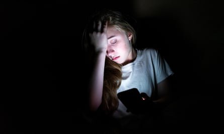 Fille bouleversée assise dans le noir tout en utilisant son smartphone. La lumière de l'écran éclaire son visage.