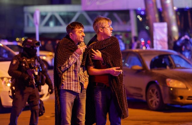 Des hommes se couvrent de couvertures tandis qu'un agent russe chargé de l'application des lois monte la garde près de la salle de concert en feu de l'hôtel de ville de Crocus, à la suite d'une fusillade signalée, à l'extérieur de Moscou, en Russie.