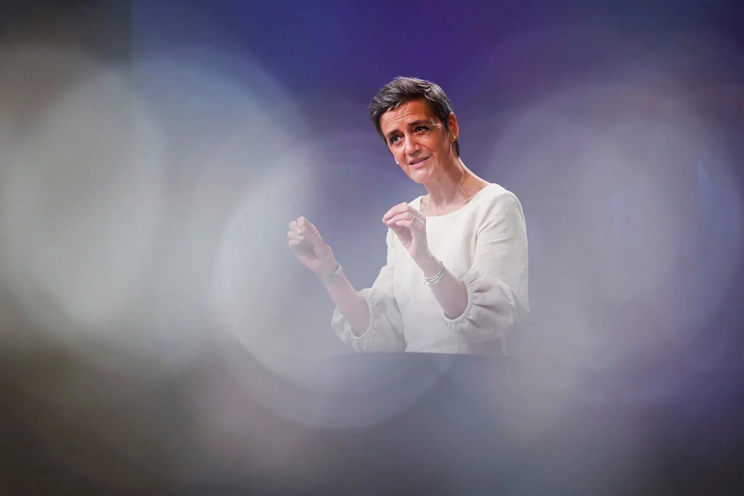 A woman with short hair wearing a white top is seen through blurred light circles as she gestures with both hands.
