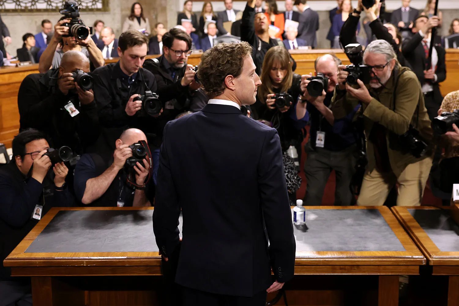Photographers swarm around a man wearing a suit with his back to the camera as he stands in front of a table to testify before members of the Senate Judiciary, seen in the distance.