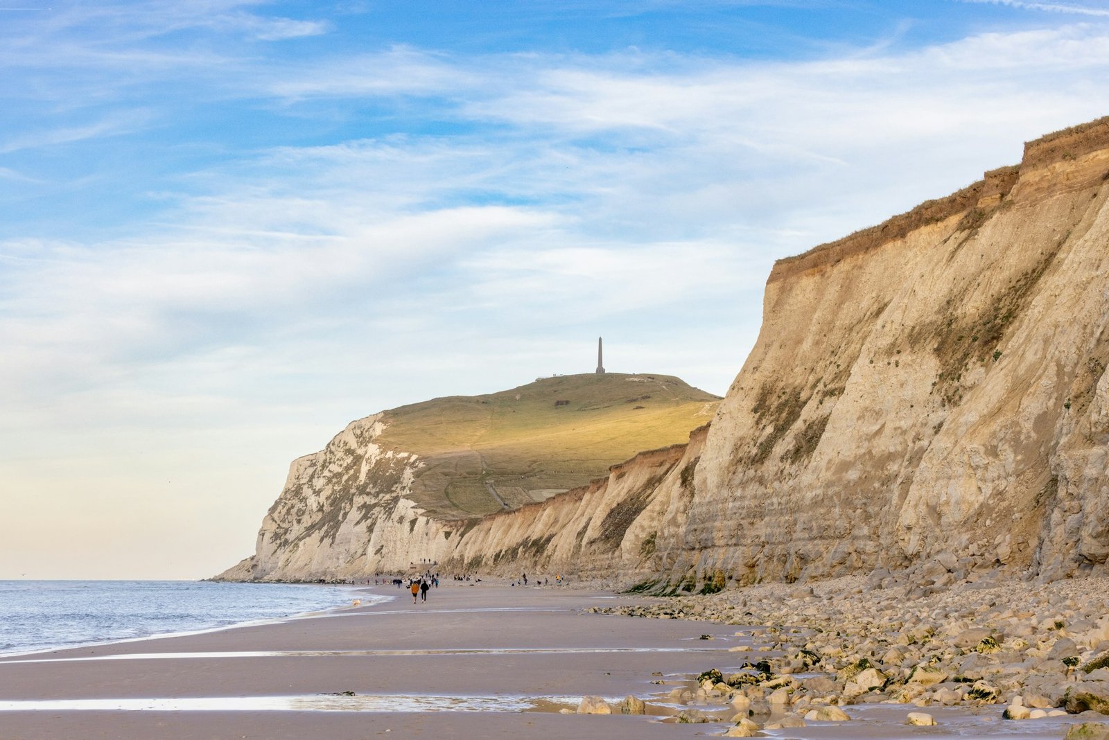 Les gens marchent le long d’une plage à côté d’une grande falaise surmontée d’un obélisque