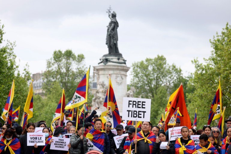 Les manifestants tibétains se rassemblent à Paris à l'arrivée de Xi Jinping.  Ils portent des pancartes indiquant « Tibet libre » et brandissent des drapeaux tibétains.  Il y a une statue derrière eux.