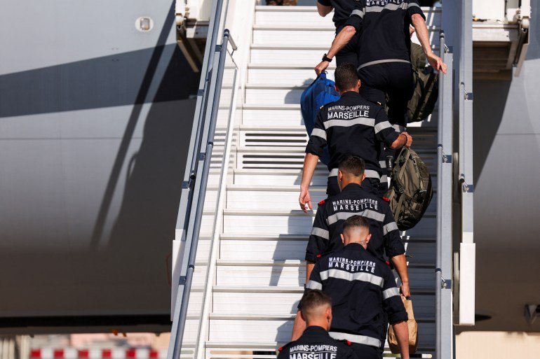 Des membres du bataillon des pompiers de la Marine de Marseille montent les marches pour monter à bord d'un avion. Ils portent des uniformes bleus avec écrit « Marins Pompiers, Marseille » au dos sur les épaules.