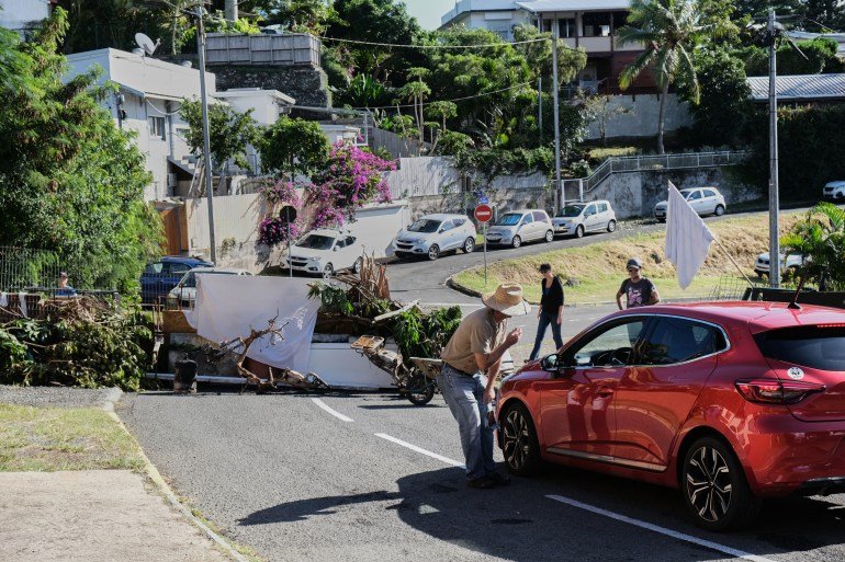 Barricade de fortune d'habitants dans un quartier de Nouméa. Un homme parle au conducteur d’une voiture. D'autres personnes sont en arrière-plan. Il y a un drapeau blanc. Il y a des maisons à flanc de colline.