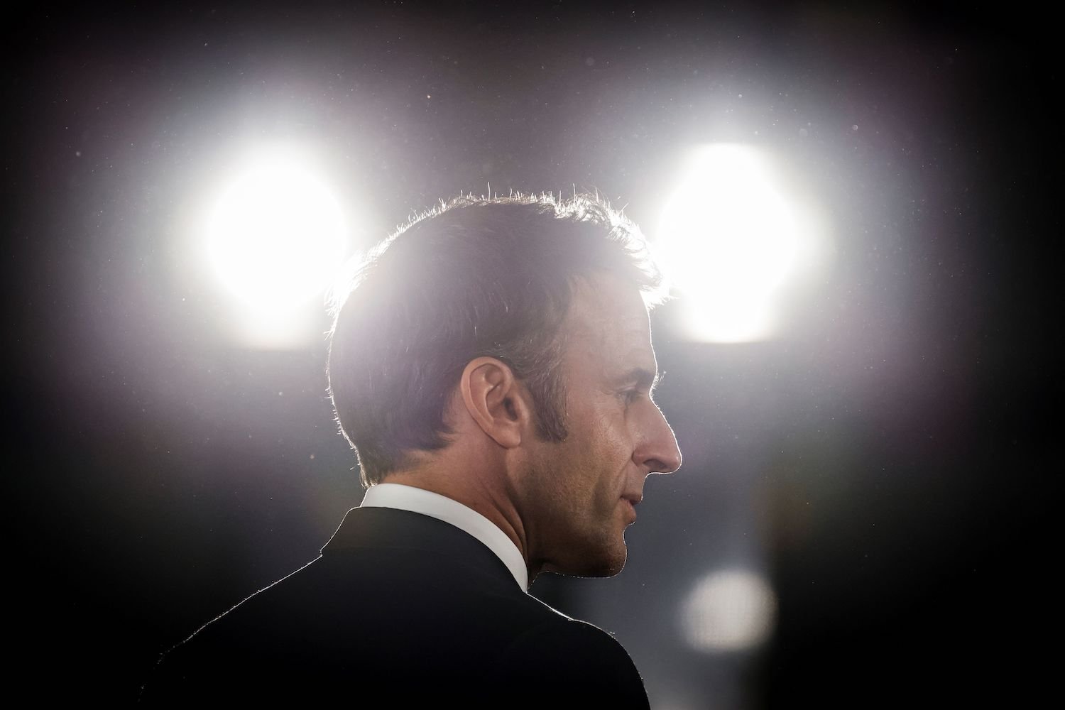 Le président français Emmanuel Macron est vu de profil lors d'un discours sur scène.  Deux lumières blanches brillantes brillent en arrière-plan, une de chaque côté de sa tête.  Macron est un homme d'une quarantaine d'années aux cheveux noirs, vêtu d'un costume noir.
