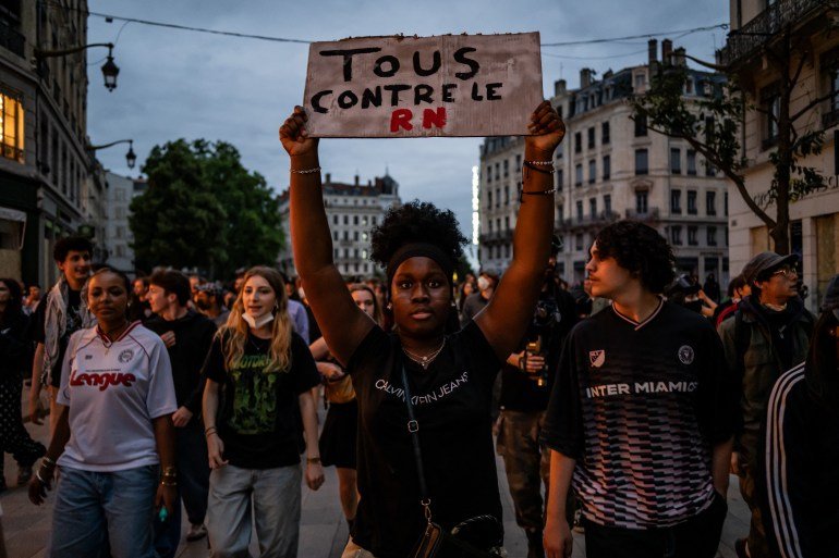 À Lyon, un manifestant brandit une pancarte indiquant « Tous contre le RN ».  Il y a des gens qui marchent derrière elle.